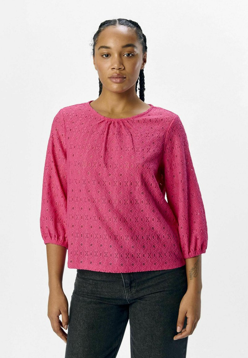 Blouse rose avec un motif œillet texturé, encolure ronde et manches trois-quarts, portée avec un jean foncé.
