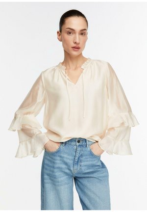 Kobieta w kremowej atłasowej bluzce z marszczonymi mankietami i niebieskich dżinsach z wysokim stanem, ręce w kieszeniach, neutralny wyraz twarzy, jednolite białe tło.