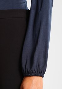 Mörkblå långärmad topp med rynkade manschetter, tillverkad i ett mjukt tyg. Svart midja syns, vilket framhäver de kontrasterande texturerna.