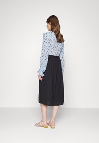 Blusa de manga longa azul com estampado floral e punhos com babados, combinada com uma saia midi preta plissada. Sandálias leves com detalhes em madeira no salto.