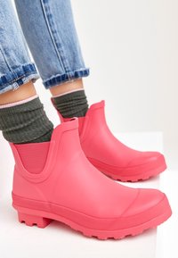 Personne portant des bottes en caoutchouc roses à hauteur de cheville avec des chaussettes vert foncé et un jean bleu clair retroussé sur un fond blanc.