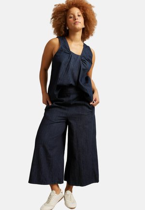 Vrouw met krullend haar draagt een mouwloze donkere denimtop en wijde cropped broek gecombineerd met witte vetersneakers.