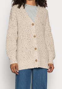 Beige stickad cardigan med V-ringning, textil med struktur och fem knappar, i kombination med lösa blå denimjeans.