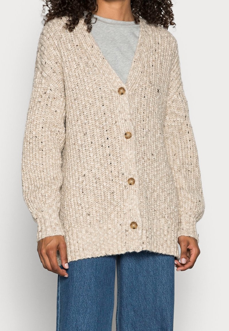 Beige stickad cardigan med V-ringning, textil med struktur och fem knappar, i kombination med lösa blå denimjeans.