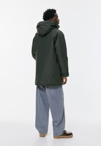 Bărbat îmbrăcat cu o haină de iarnă cu glugă de culoare verde închis, pantaloni lejeri de blugi de culoare albastră deschisă și cizme maronii, stând cu corpul întors într-o parte pe un fundal alb.