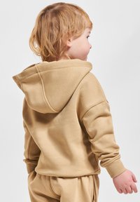 Sudadera con capucha color beige que presenta puños acanalados, textura suave y la vista trasera de un niño con el cabello a la altura de los hombros.