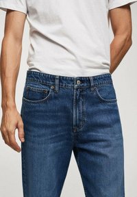 Jean en denim bleu avec un design classique à cinq poches, doté d'une fermeture éclair et d'un bouton, de coutures solides et d'une finition légèrement délavée.