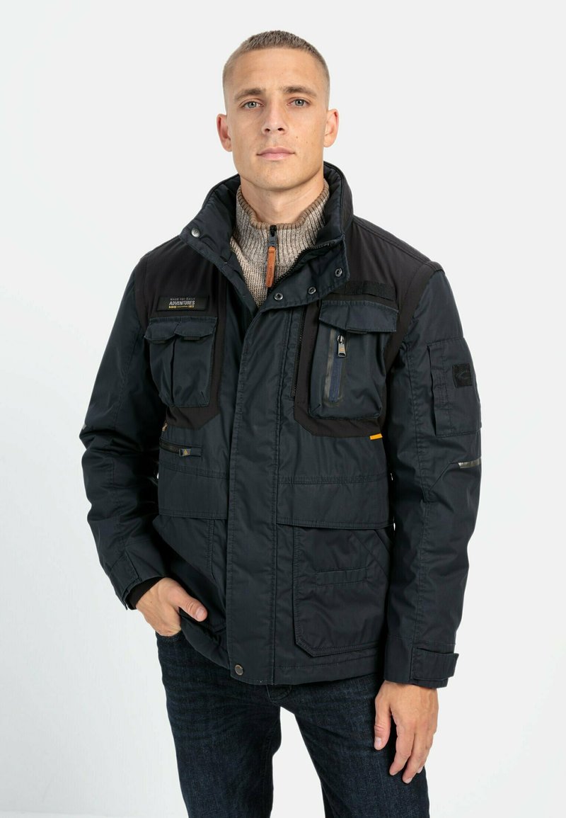 Sort udendørsjakke med flere lommer foran, høj krave og lynlåslukning. Har grå detaljer og en tekstureret indvendig foring.