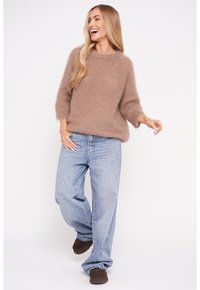 Brązowy puszysty sweter z okrągłym dekoltem, rękawami 3/4, w połączeniu z luźnymi niebieskimi dżinsami i brązowymi butami wsuwanymi.