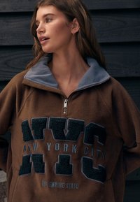 Brun fleecepullover med grå krave, prydet med præget sort tekst "NEW YORK CITY" og en lynlåslukning foran. Blød tekstur, afslappet pasform.