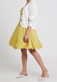 Gul blommig klänning med utsvängd kjol, tillsammans med en vit stickad cardigan med stora knappar. Vita slip-on skor med ett vävt mönster.