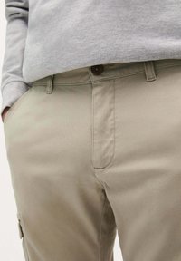 Pantaloni din bumbac bej cu croială dreaptă, prevăzuți cu închidere cu nasture, buzunare laterale și detalii de cusătură discrete. Țesătura texturată este vizibilă.
