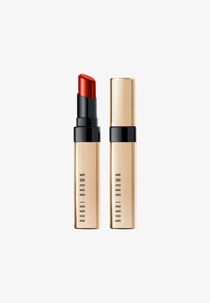 Rouge à lèvres dans un étui doré et noir. Le raisin est biseauté, et l'étui présente un branding minimaliste. Texture lisse et métallique.