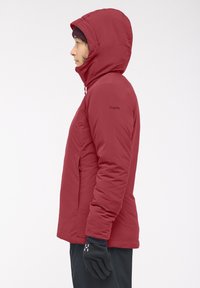 Veste isolée rouge avec capuche, poches à fermeture éclair et gants noirs. Texture de tissu lisse, design ajusté avec une forme légèrement cintrée.