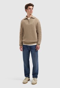 Beige, ribbeltrui met halve rits en kraag, gedragen over een wit shirt, gecombineerd met blauwe jeans en lichtgekleurde schoenen. Staande pose tegen een neutrale achtergrond.