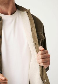 Corduroy jacket in olive green with a white fleece lining, featuring buttons and a folded collar. Worn over a plain white T-shirt.