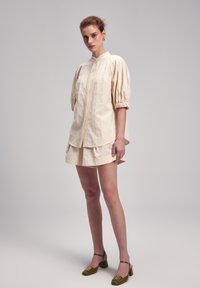 Helles beige Leinenbluse mit Puffärmeln, filigrane Stoffstruktur, kombiniert mit einem passenden Minirock und grünen High Heels.