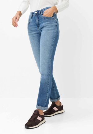 Person, die hellblaue hochgekrempelte Jeans und dunkelbraune Sneakers mit hellrosa Details trägt, steht vor einem weißen Hintergrund.