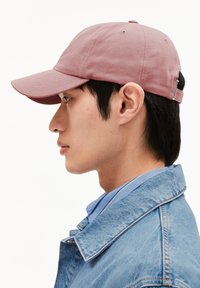 Baumwoll-Baseballmütze in staubigem Rosé, mit einem gebogenen Schild und verstellbarem Riemen. Getragen mit einer Jeansjacke und einem hellblauen Hemd mit Kragen.