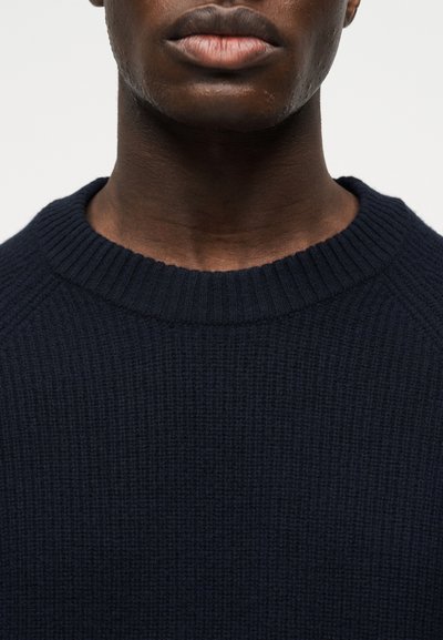 Κοντινό πλάνο του κάτω μέρους του προσώπου και του λαιμού ενός ατόμου που φοράει ένα σκουρό μπλε πουλόβερ με ριμπ ύφανση και λαιμόκοψη στρογγυλή.
