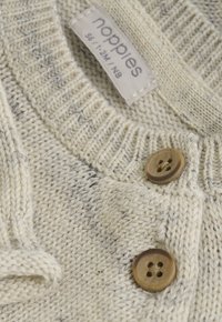 Jasnobeżowy, dziany sweterek dla niemowląt z dwoma brązowymi guzikami oraz metką z nazwą marki i rozmiarem dla dzieci w wieku 1-2 miesięcy.