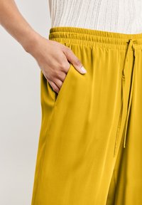 Żółte, lekkie spodnie z elastycznym pasem i boczną kieszenią. Gładka faktura z detalem sznurka przy pasie.