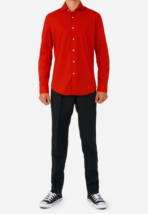 Mężczyzna ubrany w jaskrawą czerwoną koszulę na guziki, czarne spodnie i czarne niskie trampki z białymi sznurówkami, stojący na tle jednolitego tła.