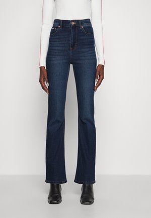 Mannequin portant un jean bleu foncé taille haute à jambe droite avec poches avant et bottines noires sur un fond uni.