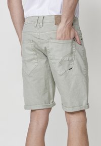 Oseba v svetlo sivih jeans kratkih hlačah z zavihanimi robovi, beli majici in z desno roko v zadnjem žepu, prikazana od pasu navzdol s hrbtne strani.