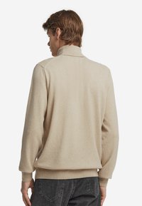 Mann in beigem Rollkragenpullover und dunkelgrauen Jeans, von hinten vor weißem Hintergrund gezeigt.