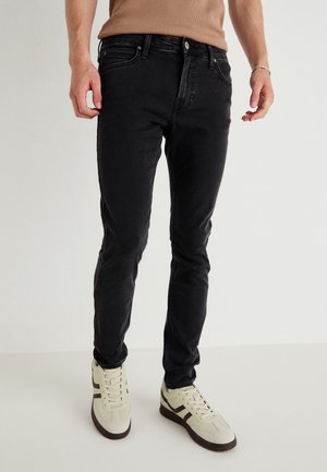 Man die strakke zwarte jeans en beige sneakers draagt, staande op een witte vloer tegen een effen achtergrond.