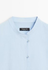 Light blue cotton shirt featuring a mandarin collar, subtle texture, and two buttons at the front for closure. Brand label visible.