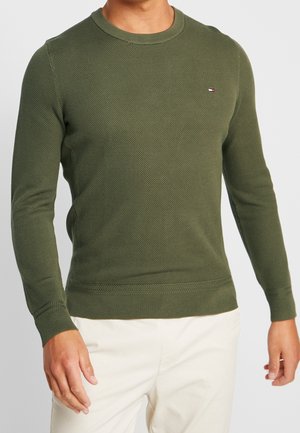 Man draagt een aansluitende olijfgroene gestructureerde trui met lange mouwen en een klein vlaglogo op de borst en crèmekleurige broek.