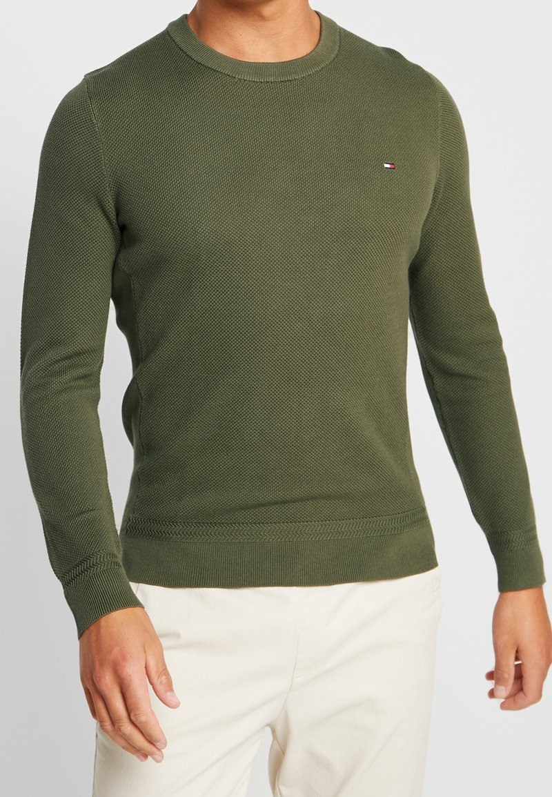 Homme portant un pull ajusté vert olive texturé à manches longues avec un petit logo drapeau sur la poitrine et un pantalon couleur crème.