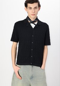 Chemise en tricot noire à manches courtes avec un motif de vagues texturé, fermée par des boutons, avec un col. Portee avec un sous-vêtement blanc et un short en jean.