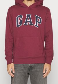 Sweat à capuche bordeaux avec de grandes lettres "GAP" bleu marine et blanches sur la poitrine, porté par une personne portant un jean gris, debout les bras détendus.