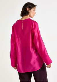 Lys rosa, langærmet skjorte med dyb rygåbning, rundet kant og glat tekstur, parret med mørkfarget bukser.
