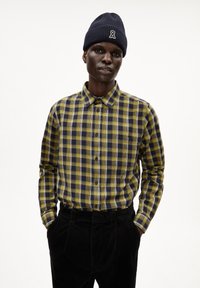 Geverfd shirt in geel en marineblauw, knoopsluiting, katoen stof, lange mouwen. G gedragen met zwarte corduroy broek en een marineblauwe beanie.