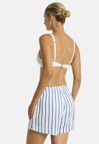 Kobieta z upięciem, odwrócona plecami, w pasiakowej biustonoszowej bluzce i szortach w niebieskie paski, na jednolitym białym tle.