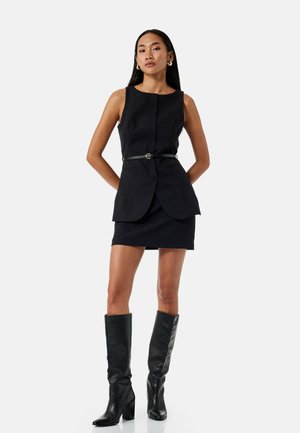 Schwarzes tailliertes Kleid mit einem gefalteten Design, das über einen taillierten Gürtel und zwei vordere Taschen verfügt, kombiniert mit kniehohen schwarzen Stiefeletten mit Absatz.
