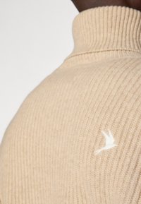 Beige geribbelde trui met hoge hals en wit vogellogo op de schouder. Heeft een zachte textuur en een getailleerde pasvorm.