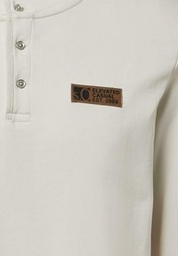 Crème-kleurige geribde henley-shirt met een drie-knopen sluiting en een bruine leren patch met het label "s.o Elevated Casual Est. 1969."