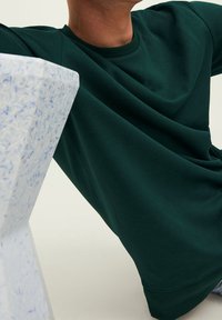 Felpa verde scuro realizzata in tessuto liscio con scollatura rotonda classica, dotata di polsini e orlo a coste, abbinata a una superficie bianca testurizzata.