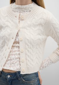 Cable knit vest in licht beige, met een geribbelde kraag en manchetten, zes lichte knopen, gedragen over een gestructureerd wit topje.