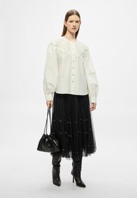 Biela blúzka s vyšívaným detailom na ramene, v kombinácii s čiernou tylovou sukňou ozdobenou perlami, čiernou kabelkou a čiernymi čižmami po kolená.