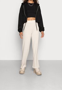Mujer vistiendo un hoodie negro recortado, un top negro corto, pantalones anchos color crema y zapatillas gruesas beige frente a un fondo claro y liso.