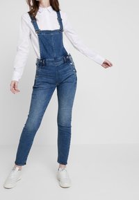 Denimbyxor med justerbara axelband, framsida med ficka och sidoknappar. Bärs över en vit skjorta med krage, åtsittande passform, i medelblå tvätt.