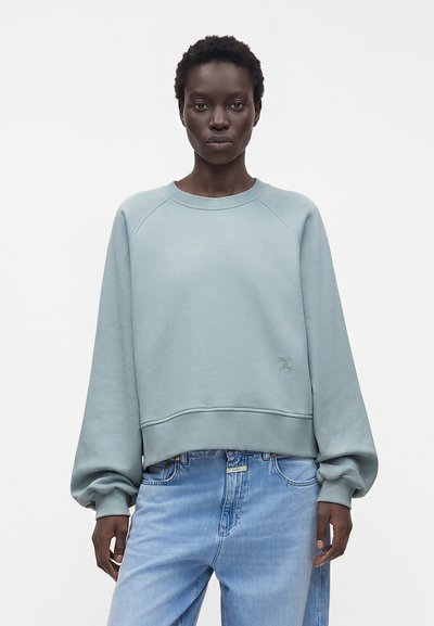 Femme aux cheveux courts portant un sweat-shirt ample bleu clair et un jean bleu clair sur un fond uni.