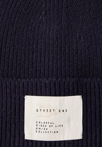 Stickad mössa i mörk marinblå, med ribbat mönster, och en vävd etikett med "Street One" och kollektionsdetaljer i gräddvit och svart.