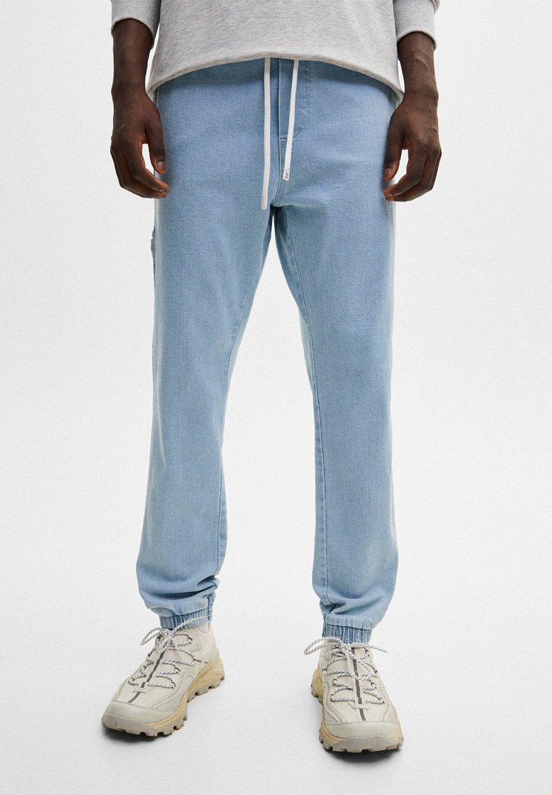 Joggers bleu clair en tissu doux, avec poignets élastiques, taille à cordon et poches latérales, assortis à des baskets beiges.